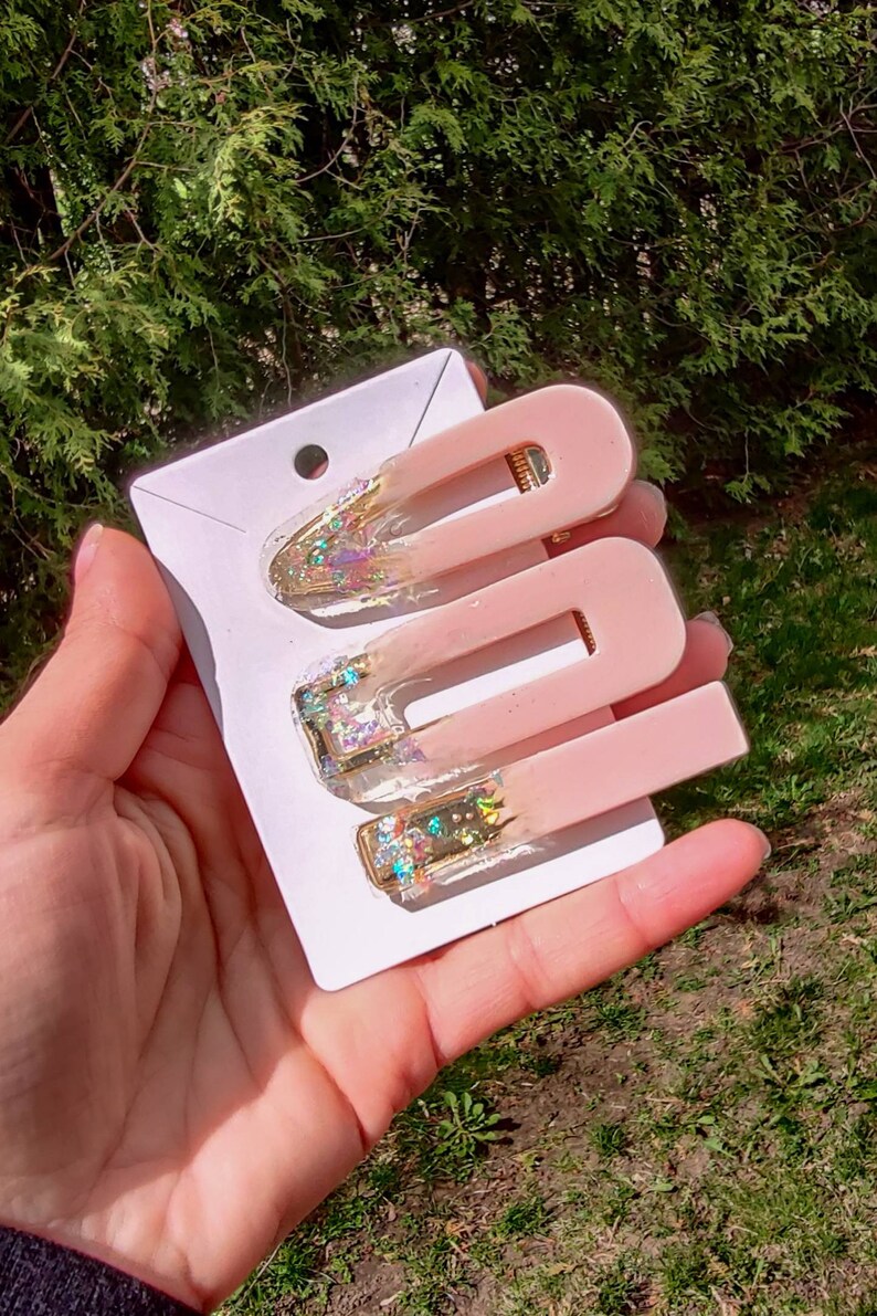 May include: Three pink hair clips with gold glitter accents. The clips are on a white card.