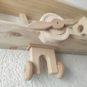 May include: Wooden airplane shelf with two shelves. The airplane is made of natural wood and has a propeller. The shelf is mounted on a wall.