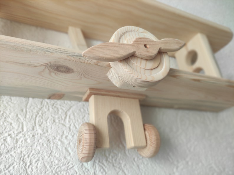 May include: Wooden airplane shelf with two shelves. The airplane is made of natural wood and has a propeller. The shelf is mounted on a wall.