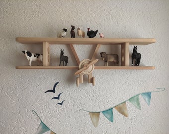 May include: Wooden airplane shelf with various animal figurines, including a cow, sheep, a horse, and a turkey. The shelf is mounted on a white wall with a blue and green garland hanging below.
