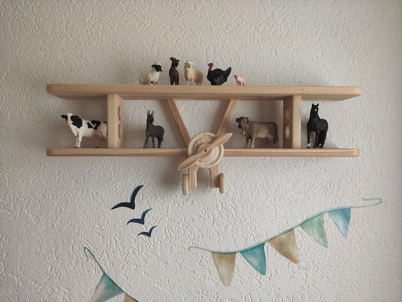 May include: Wooden airplane shelf with various animal figurines, including a cow, sheep, a horse, and a turkey. The shelf is mounted on a white wall with a blue and green garland hanging below.