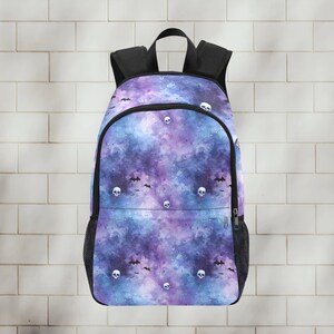May include: A black backpack with a galaxy print featuring white skulls and bats. The background is a watercolor-style galaxy with shades of purple, blue, and pink.