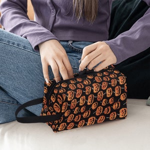 May include: A black zippered makeup bag with a repeating pattern of orange jack-o'-lantern faces on a black background.