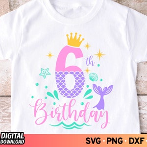 Peut inclure: Un t-shirt blanc avec un numéro six en forme de queue de sirène rose et violette, une couronne dorée et le texte "6th Birthday".