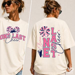 Op de afbeelding: Een off-white t-shirt met korte mouwen en een grafisch ontwerp met roze en marineblauwe tekst die One Last Cheer leest. Het ontwerp bevat illustraties van pompons en een megafoon, met de naam Olivia geïntegreerd in de print op de rug.