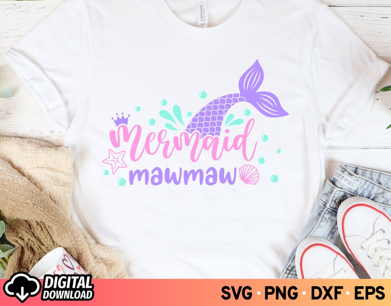 May include: A white t-shirt with a purple mermaid tail, pink crown, and the text "Mermaid Mawmaw" in pink and purple lettering. The design is surrounded by blue and green bubbles.