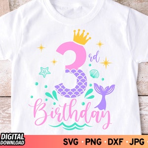 May include: A white t-shirt with a pink and purple mermaid-themed design. The design includes a number "3" with a crown, a mermaid tail, and the text "3rd Birthday".