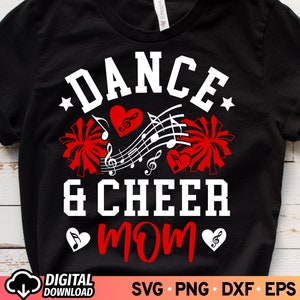May include: Black t-shirt with white text that reads "Dance & Cheer Mom" with a red heart and musical notes. There are two white pom-poms on either side of the text.