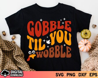 Może przedstawiać: Czarny T-shirt z grafiką w stylu retro, na której widnieje napis "Gobble Til You Wobble" w kolorze pomarańczowym, czerwonym i żółtym. Grafika zawiera również głowę indyka i liść.