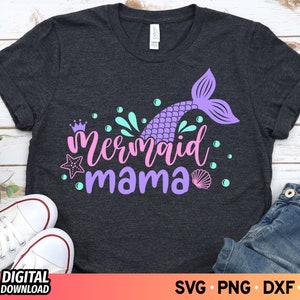 May include: A dark gray t-shirt with a purple mermaid tail and the text "mermaid mama" in pink and purple lettering. The design also includes a crown, starfish, seashells, and bubbles.