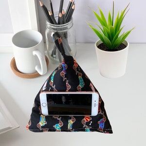 K&ouml;nnte beinhalten: Ein schwarzer Telefonst&auml;nder mit einem Dackel-Muster. Der St&auml;nder ist aus Stoff und hat eine Tasche zum Halten eines Telefons. Der St&auml;nder steht auf einer wei&szlig;en Oberfl&auml;che mit einer wei&szlig;en Tasse und einer Topfpflanze.