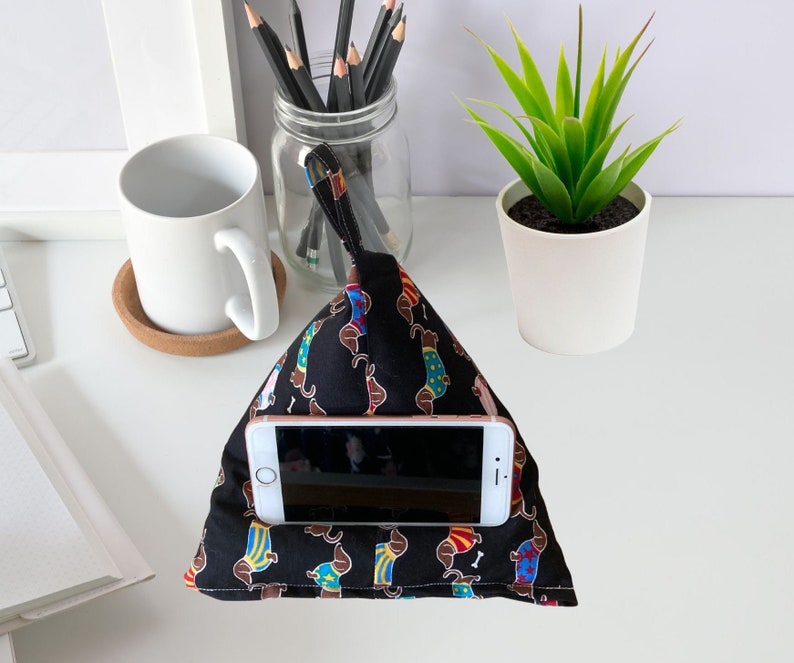 K&ouml;nnte beinhalten: Ein schwarzer Telefonst&auml;nder mit einem Dackel-Muster. Der St&auml;nder ist aus Stoff und hat eine Tasche zum Halten eines Telefons. Der St&auml;nder steht auf einer wei&szlig;en Oberfl&auml;che mit einer wei&szlig;en Tasse und einer Topfpflanze.