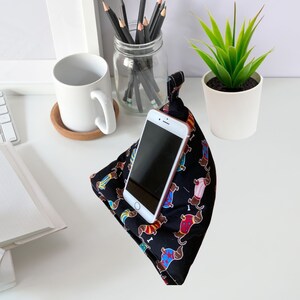 K&ouml;nnte beinhalten: Ein schwarzer Telefonst&auml;nder mit einem Dackel-Muster. Der St&auml;nder ist aus Stoff und hat eine Tasche zum Halten eines Telefons. Der St&auml;nder steht auf einer wei&szlig;en Oberfl&auml;che mit einer wei&szlig;en Tasse und einem Glas mit Bleistiften.