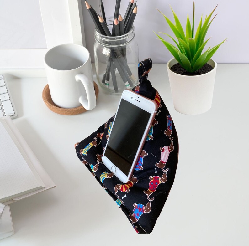 K&ouml;nnte beinhalten: Ein schwarzer Telefonst&auml;nder mit einem Dackel-Muster. Der St&auml;nder ist aus Stoff und hat eine Tasche zum Halten eines Telefons. Der St&auml;nder steht auf einer wei&szlig;en Oberfl&auml;che mit einer wei&szlig;en Tasse und einem Glas mit Bleistiften.