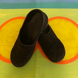 May include: A pair of black suede clogs with a rounded toe. The clogs are on a colorful background.