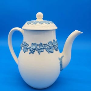 May include: White ceramic teapot with a blue grape vine design around the body and lid. The teapot has a curved spout and a handle. The background is a solid blue.