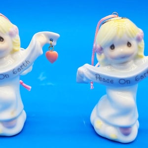 May include: Two ceramic angel figurines with blonde hair and white robes. Each angel holds a banner that reads "Peace On Earth" and a red heart. The figurines have a pink ribbon for hanging and are on a blue background.