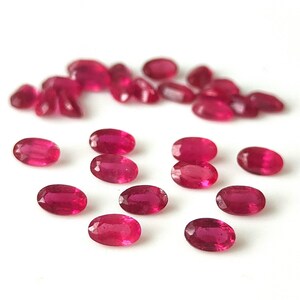 Może przedstawiać: Kolekcja owalnych, ciemnoczerwonych kamieni szlachetnych. Wypolerowane rubiny są rozrzucone na białej powierzchni, o różnych rozmiarach i błyszczącym, półprzezroczystym wyglądzie. Te kamienie szlachetne nadają się do tworzenia biżuterii.