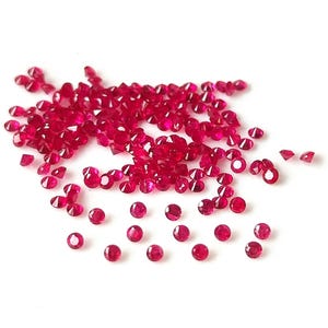 Può includere: Una dispersione di piccole gemme rotonde, sfaccettate, di colore rosso intenso. Le gemme sono sparse su una superficie bianca, alcune raggruppate e altre distanziate. Le gemme hanno una qualità brillante e riflettente.