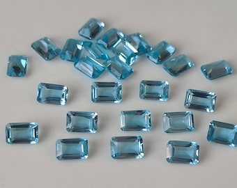 May include: A collection of rectangular, light blue gemstones with a polished finish. The gems are scattered across a white surface, showcasing their clarity and color. The gemstones are of various sizes and shapes.