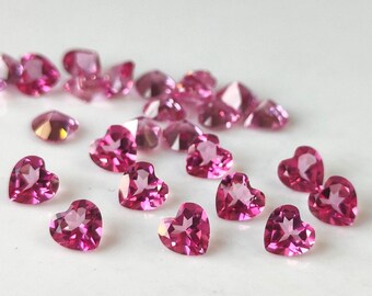May include: A collection of faceted, heart-shaped gemstones in varying sizes. The gems are a vibrant pink color and are scattered across a white surface. The light catches the facets, creating a sparkling effect.