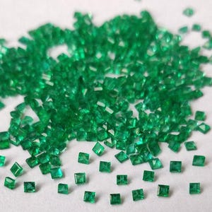 May include: A scattering of small, square-cut emerald gemstones on a white surface. The gemstones are a vibrant green colour and appear faceted, catching the light. The image is a close-up, showcasing the details of the gems.