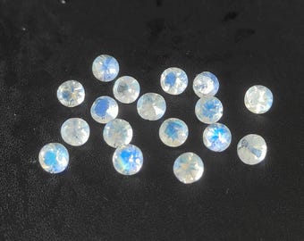 May include: A collection of round, iridescent moonstone gemstones scattered on a dark surface. The stones display a range of colors, including blue, white, and hints of yellow, with a polished, faceted appearance. These gemstones are ideal for jewelry making.
