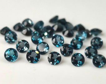 Op de afbeelding: Een verzameling ronde, gefacetteerde edelstenen in tinten teal en donkerblauw. De edelstenen zijn verspreid op een wit oppervlak, met licht dat weerkaatst op hun gepolijste oppervlakken. De edelstenen variëren enigszins in grootte en vorm.