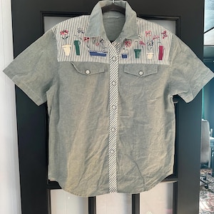 Op de afbeelding: Een shirt met korte mouwen en knoopsluiting in een gedempte saliegroene stof. Het bovenste gedeelte van de borst heeft een wit en grijs gestreept paneel met geborduurde kleurrijke potbloemen. Het bevat twee borstzakken met kleppen en een gestreepte knoopsluiting.