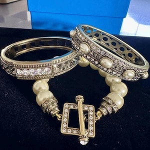 Könnte beinhalten: Zwei goldfarbene Armbänder mit klaren und perlenartigen Verzierungen. Eines ist ein Scharnierarmreif mit klaren Steinen. Das andere ist ein Scharnierarmreif mit perlenartigen Steinen. Die Armbänder sind durch eine perlenartige Kette und einen quadratischen Verschluss verbunden.