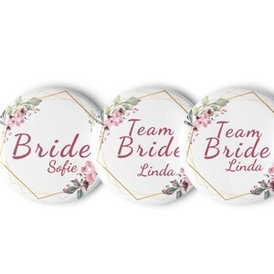 Könnte beinhalten: Fünf runde weiße Knöpfe mit goldener Umrandung und Blumendesigns. Die Knöpfe tragen den Text "Team Bride" mit einem Namen unter jedem. Die Namen sind Sofie, Linda, Linda, Lisa-Marie und Daisy.