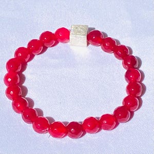 Könnte beinhalten: Ein Armband aus roten Edelsteinen mit einer silbernen quadratischen Perle. Das Armband besteht aus runden Perlen und ist etwa 18 cm lang.