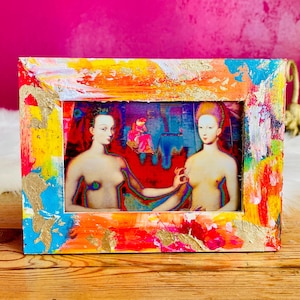 Op de afbeelding: Een kleurrijke, handgeschilderde fotolijst met een levendig abstract ontwerp in tinten rood, oranje, geel, blauw en goud. Het frame omringt een afdruk van twee naakte figuren. Het kunstwerk wordt op een houten oppervlak weergegeven.