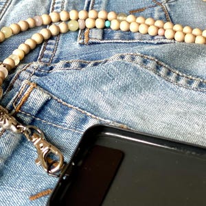 May include: A wooden beaded phone charm with a silver clasp and turquoise and pink beads. The charm is lying on a blue denim background.