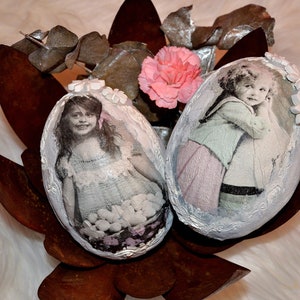 May include: Two white painted Easter eggs decorated with vintage photos of children and lace trim. The eggs are nestled in a rusty metal flower pot with a pink carnation.