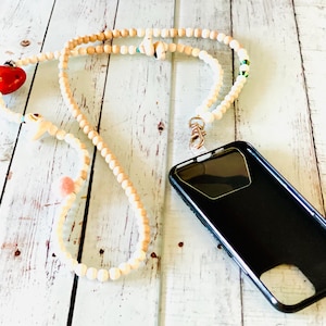 May include: A white wooden beaded phone strap with a red heart charm, a white shell charm, and a green and white bead. The strap is attached to a black phone case.