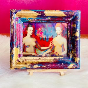 Op de afbeelding: Een ingelijste print met twee figuren in een kleurrijke, abstracte stijl. De lijst is goudkleurig, blauw en paars met een houten ezel. Het kunstwerk heeft een rode en blauwe achtergrond.