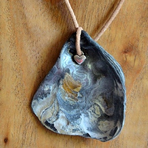 Puede incluir: Un collar con un gran colgante de concha de mar gris oscuro y blanco con un dije de corazón plateado. El colgante tiene una textura natural y orgánica.