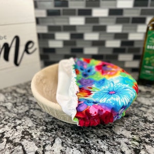 May include: A white bowl with a colorful floral fabric cover. The fabric has a variety of flowers in shades of pink, blue, green, and yellow. The bowl is sitting on a black and white countertop.