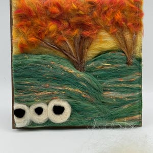 May include: A fibre art landscape scene featuring trees with orange and yellow foliage, set against a yellow sky and green hills. The artwork is framed in a dark brown wooden frame, with three white and black circular shapes at the bottom.