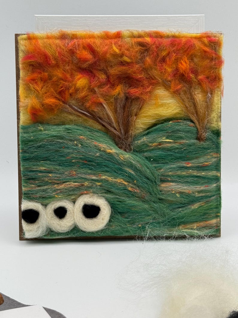 May include: A fibre art landscape scene featuring trees with orange and yellow foliage, set against a yellow sky and green hills. The artwork is framed in a dark brown wooden frame, with three white and black circular shapes at the bottom.