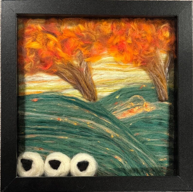 May include: Framed fibre art landscape. Two trees with orange and red leaves stand over green hills and a yellow and orange sky. Three white and black objects are at the bottom of the artwork.