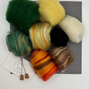 May include: A selection of vibrant wool roving in green, yellow, orange, and brown, displayed on a grey felt square. A small card with the text "Ewes and Co" and two cork stoppers are also included. Suitable for craft projects.