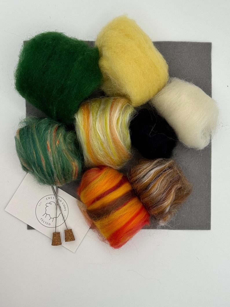 May include: A selection of vibrant wool roving in green, yellow, orange, and brown, displayed on a grey felt square. A small card with the text "Ewes and Co" and two cork stoppers are also included. Suitable for craft projects.