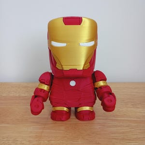 Peut inclure: Figurine d'Iron Man rouge et or. La figurine a un casque dor&eacute; avec des fentes oculaires blanches, un corps rouge et des accents dor&eacute;s sur les bras et les jambes. La figurine est pos&eacute;e sur une surface en bois.