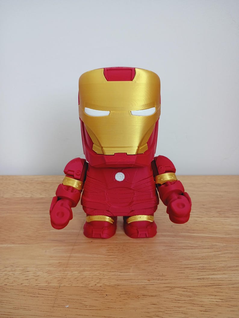 Peut inclure: Figurine d'Iron Man rouge et or. La figurine a un casque dor&eacute; avec des fentes oculaires blanches, un corps rouge et des accents dor&eacute;s sur les bras et les jambes. La figurine est pos&eacute;e sur une surface en bois.