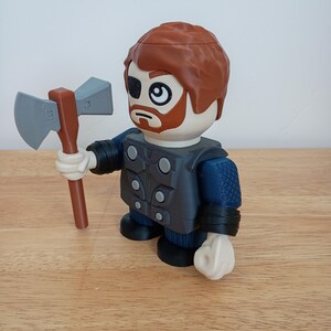 Peut inclure: Une figurine de jouet repr&eacute;sentant un personnage aux cheveux roux, une barbe et un cache-&oelig;il, tenant une hache grise et marron. La figurine porte une tenue bleue et grise avec des gants et des bottes noirs. Le jouet est sur une surface en bois.