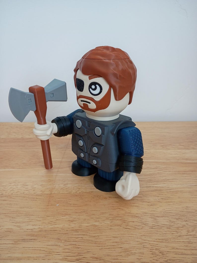 Peut inclure: Une figurine de jouet repr&eacute;sentant un personnage aux cheveux roux, une barbe et un cache-&oelig;il, tenant une hache grise et marron. La figurine porte une tenue bleue et grise avec des gants et des bottes noirs. Le jouet est sur une surface en bois.