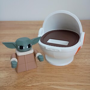 May include: A green and tan Grogu figure with large ears and a tan robe stands next to a white and brown pod. The pod has an open top and a circular base. The figure is a toy or collectible item.