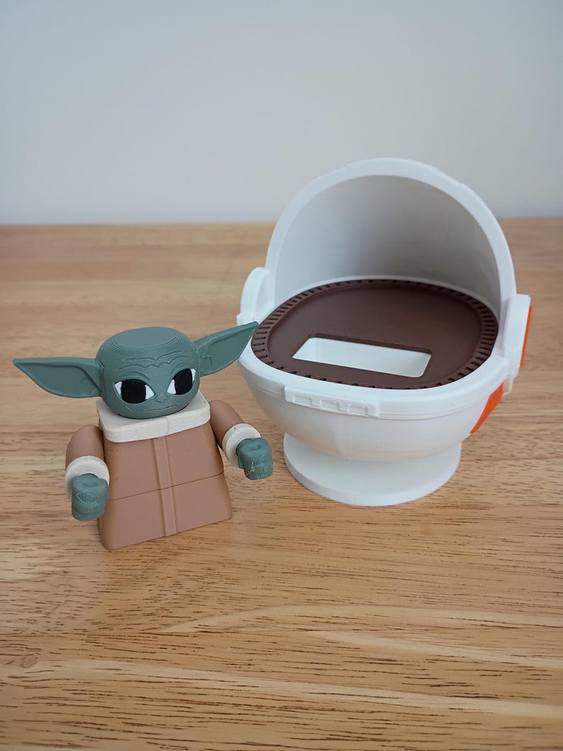 May include: A green and tan Grogu figure with large ears and a tan robe stands next to a white and brown pod. The pod has an open top and a circular base. The figure is a toy or collectible item.